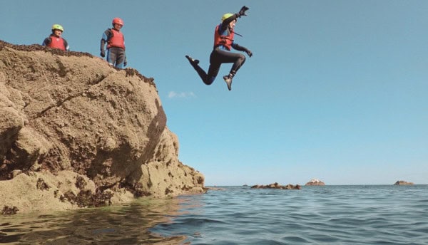 Three people wearing helmets and life jackets, with one person jumping off a rock into the sea.